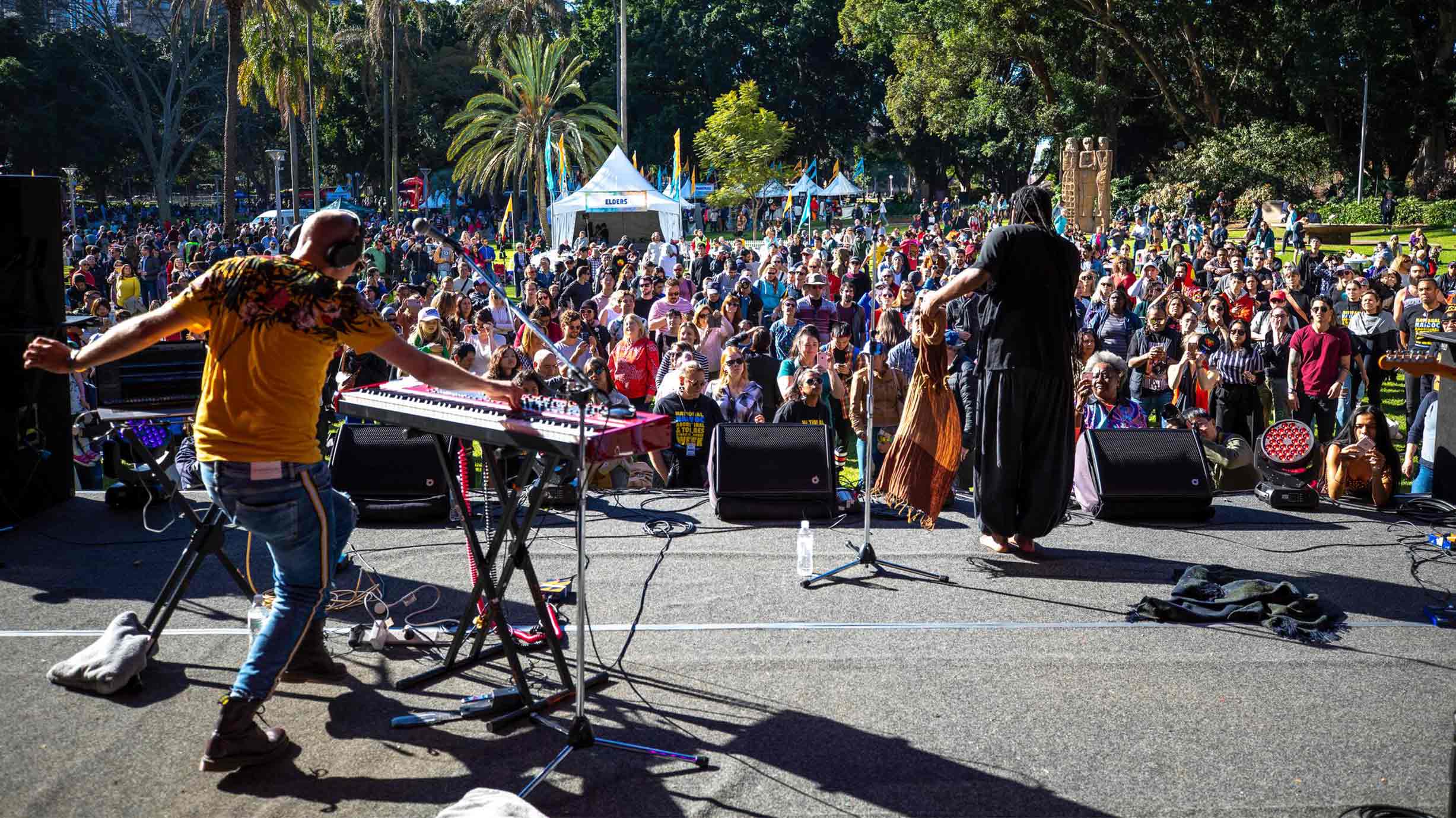 2 performers on a stage in a park in front of a large crowd of people